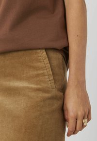 Jupe en velours côtelé brun avec des côtes verticales, dotée de poches latérales. Une main portant une bague en or est partiellement visible, mettant en valeur la texture du tissu.