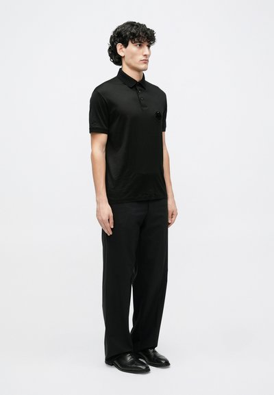 Juoda polo marškinėliai iš lygios medžiagos, su sagų siuvimo juostele ir mažu apvaliu logotipu kairėje krūtinės pusėje. Dėvimi su laisvomis juodomis kelnėmis ir batais.