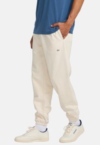 Hellbeige Sweatpants mit einem gerippten Bund und Seitentaschen, kombiniert mit einem blauen Shirt und weißen Sneakers.