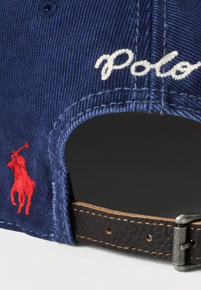 Tamsiai mėlynas medvilnės beisbolo kepuraitė su balta "Polo" siuvinėjimu ir raudonu arkliu logotipu. Ruda odinė reguliuojama juosta su sidabro akcentais.