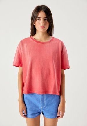 Giovane donna con capelli castani lisci indossa una maglietta corallo a maniche corte larga e pantaloncini di jeans blu, in piedi davanti a uno sfondo chiaro e uniforme.