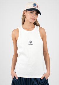 Femme portant un débardeur blanc avec logo, pantalon rayé bleu marine et casquette de baseball crème avec motif rouge et bleu. Mains dans les poches.