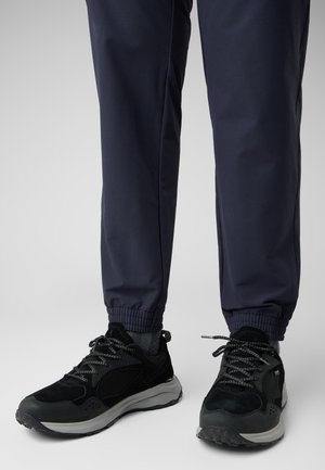 Scarpe da ginnastica nere con tomaia in rete e camoscio, dettagli riflettenti e suola grigia, abbinate a pantaloni blu con polsini elasticizzati.