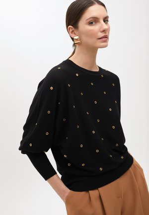 Maglione nero con dettagli in occhiali dorati, maniche lunghe e vestibilità rilassata. Abbinato a pantaloni beige a vita alta. Texture liscia.