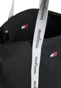 Μαύρη υφασμάτινη τσάντα ώμου, με εσωτερική τσέπη με φερμουάρ, λευκές λουρίδες με το λογότυπο και κόκκινα, λευκά και μπλε διακοσμητικά με το brand.