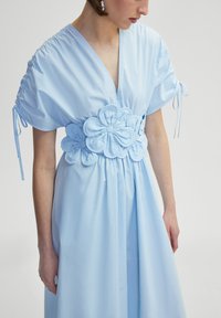 Robe bleu clair avec un décolleté en V, des manches courtes et des accents froncés. Elle présente une ceinture florale texturée et une jupe fluide. Tissu lisse.