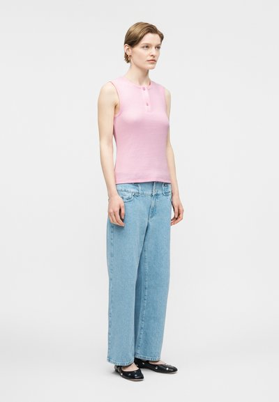 Γυναίκα που στέκεται, φορώντας ρόζ αμάνικη μπλούζα με κουμπιά, άνετο ανοιχτό μπλε τζιν και μαύρα μπαλαρίνες με ασημένιες διακοσμήσεις.