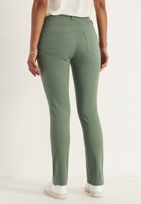 Vihreät farkut, joissa on kapea istuvuus, taka-taskut, joissa on ompeluyksityiskohtia, ja ne on yhdistetty valkoisiin tenniskenkiin. Yksinkertainen tausta.