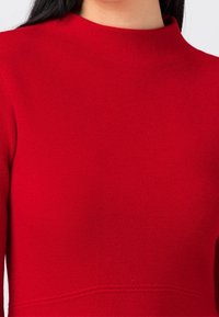 Blouse en tricot rouge avec un col haut, des manches longues et une texture lisse. Une couture détail traverse horizontalement la taille.