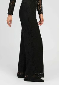 Femme portant un pantalon large en dentelle noire et un haut en dentelle transparent à manches longues, se tenant contre un fond clair et uni.