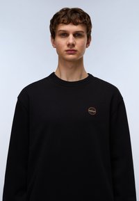 Black sweatshirt featuring a round logo patch on the left chest, ribbed crew neck, and long sleeves, made from soft cotton fabric.