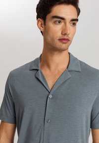 Košile s krátkým rukávem v světle modré barvě, s jemnou texturou, uvolněným límcem a dvěma knoflíky vpředu.