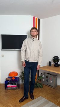 Giacca beige chiaro con colletto alto, jeans blu e scarpe scure. Indossato in una stanza con un televisore, una lampada arancione e una pianta.