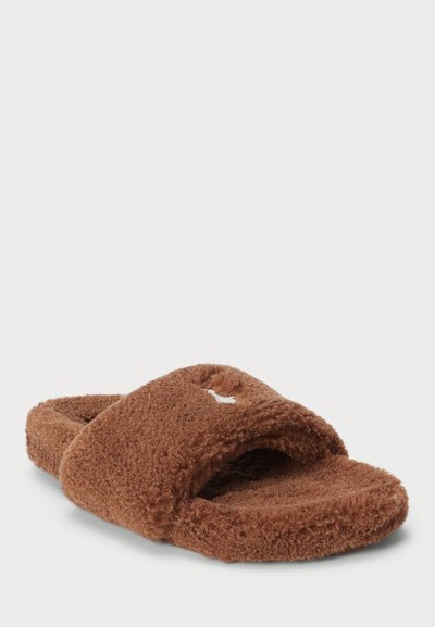 Smeđe fluffy natikače sa križnim trakama, meka tekstura i bijeli logotip kao naglasak. Otvoreni prsti s uloškom za udobnost.