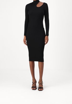 Robe midi noire côtelée avec manches longues et découpe asymétrique au niveau du décolleté, portée avec des sandales noires à talons à brides.