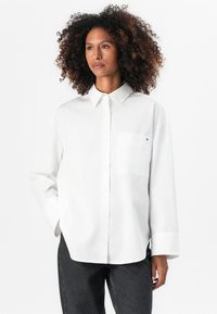 Camisa de algodón blanca con collar abotonado, mangas largas y un solo bolsillo en el pecho. Presenta un pequeño logo de la marca en el bolsillo.