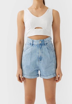 Vrouw draagt een witte, mouwloze crop top met een uitsnede aan de voorkant en high waist lichtblauwe opgerolde spijkershorts, staande tegen een egale achtergrond.