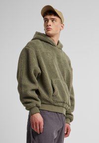 Fleece-Hoodie in olivgrün mit einer Vordertasche, gerippten Bündchen und Saum. Die Textur ist weich und leicht flauschig.