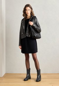 Black leather motorcycle jacket over a black dress, paired with ankle boots and a textured black bag. Model stands on a wooden floor.
