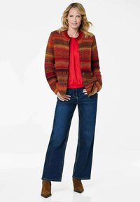 Mehrfarbige Strickjacke mit Knöpfen, die über einem roten Oberteil getragen wird. Dunkelblaue, weit geschnittene Jeans mit Steppdetails, kombiniert mit braunen Stiefeletten mit Absatz.