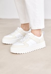 Sneakers bianchi e crema con suola testurizzata, caratterizzati da dettagli perforati, chiusura con lacci e collo imbottito per il comfort.