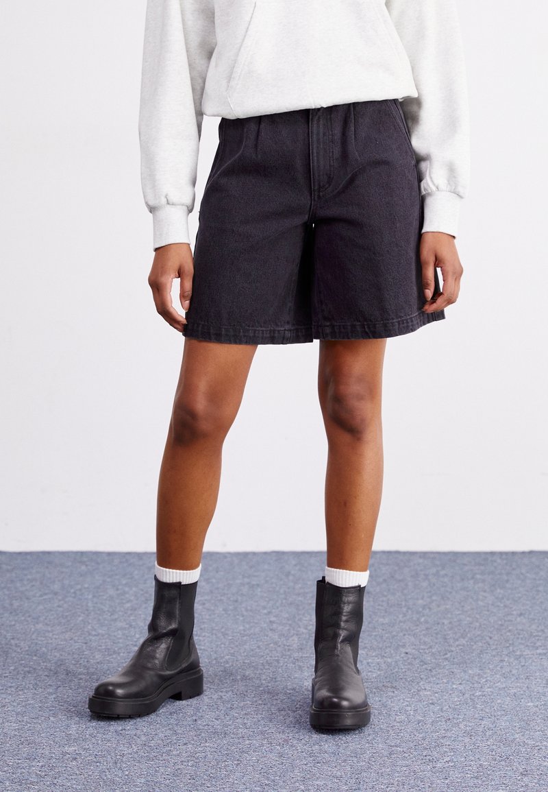 Osoba stojící na šedém koberci, oblečená v černých džínových krátkých kalhotách, bílých ponožkách, černých kožených kotníkových botách a světle šedé mikině.