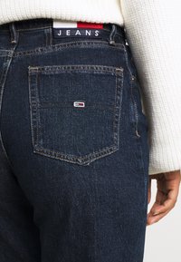 Mörkblå denimjeans med kontrasterande sömmar, försedd med en bakficka och en märkestik i midjan med "JEANS" i fet stil på en etikett.