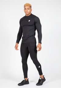 Zwarte sportieve lange mouwen top met een halve rits, gecombineerd met zwarte legging. Beide hebben een aansluitend ontwerp. Zwarte sneakers maken de outfit compleet.