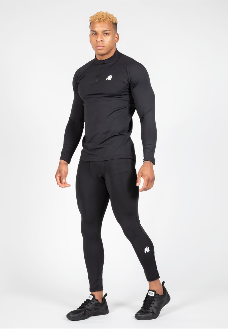 Zwarte sportieve lange mouwen top met een halve rits, gecombineerd met zwarte legging. Beide hebben een aansluitend ontwerp. Zwarte sneakers maken de outfit compleet.