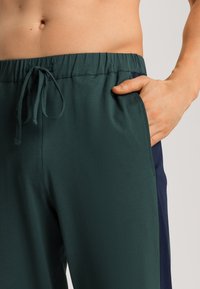 Groene broek met een elastische tailleband en trekkoord, met zijzakken en een marineblauw accent bij de zijnaad. Gladde textuur.