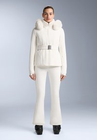 Jachetă albă cu fermoar, cu accente de blană pe glugă, talie cu curea și design cu nervuri; acompaniată de pantaloni albați ajustați și încălțăminte neagră.