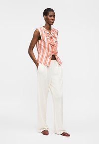 Γυναίκα με αμάνικο πορτοκαλί-λευκό ριγέ μπλουζάκι με φιόγκους, λευκό φαρδύ παντελόνι και καφέ επίπεδα παπούτσια, στέκεται με τα χέρια στις τσέπες.