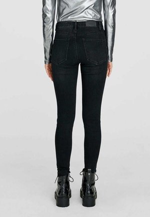 Jeans skinny de tiro alto en negro con dos bolsillos traseros, que presentan un corte ajustado y detalles de costura sutiles. Combinados con botas negras de cordones.