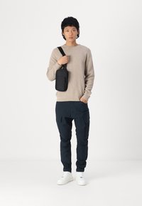 Pulover tricotat bej, pantaloni cargo negri cu buzunare laterale, și adidași albi. Geantă crossbody neagră purtată pe umăr.