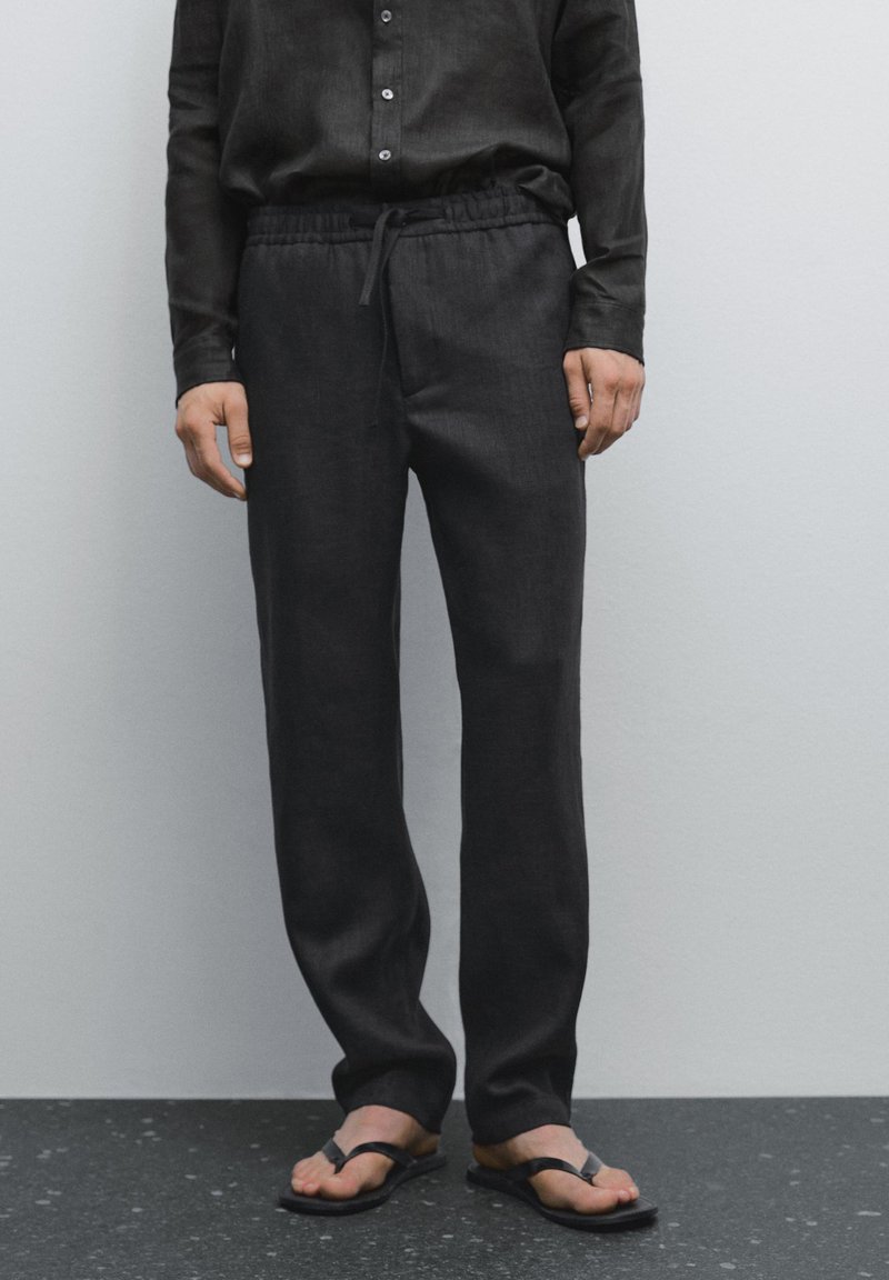 Donkere linnen broeken met een elastische tailleband, trekkoorddetail en een recht model. G gedragen met zwarte teenslippers.