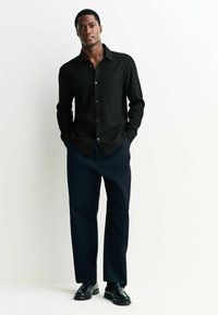 Melns ribots krekls ar norādītu apkakli, pogu aizdare priekšā; savienots ar tumši ziliem biksēm un melniem mokasīniem, stāvot ar rokām kabatās.