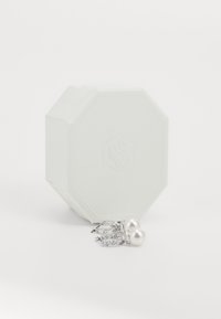 Caja de joyería octagonal blanca con una superficie texturizada. Dos pendientes de perla con herrajes de plata y acentos brillantes exhibidos al frente.