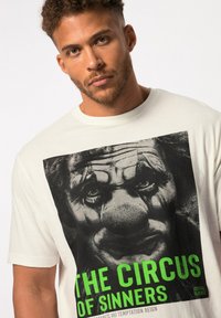 Camiseta de color crema claro con un gran gráfico de payaso en blanco y negro. El texto en la camiseta dice "EL CIRCUS DE LOS PECADORES" en negrita verde.