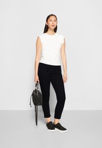 Top branco com nervuras e mangas com babados, combinado com calças pretas justas e ténis pretos. Inclui uma pequena mala tipo balde preta. Design minimalista.