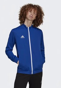 Blue zip-up jacket made of smooth fabric; features white zipper and small white logo on the left chest; stands with hands in pockets.