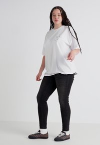 Bílá oversized tričko s jednoduchou černou grafikou, spárované s černými úzkými džíny a hnědými sportovními botami. Hladká tkanina, uvolněný střih.