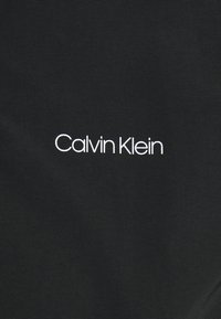Czarna tkanina z gładką, teksturowaną powierzchnią i białym logo z napisem "Calvin Klein" w nowoczesnej, bezszeryfowej czcionce.