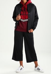 Svart bomberjacka över en vinröd hoodie med vita ränder, kombinerat med svarta cropped-byxor och vita sneakers med svarta detaljer.