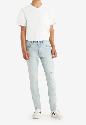 T-shirt blanc à manches courtes avec une poche, associé à un jean bleu clair légèrement déchiré et des baskets blanches avec des accents noirs.