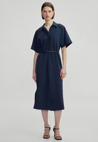 Abito blu navy con cintura, colletto, maniche corte e taglio dritto. Il tessuto appare liscio e presenta delle fessure laterali per facilitare i movimenti.