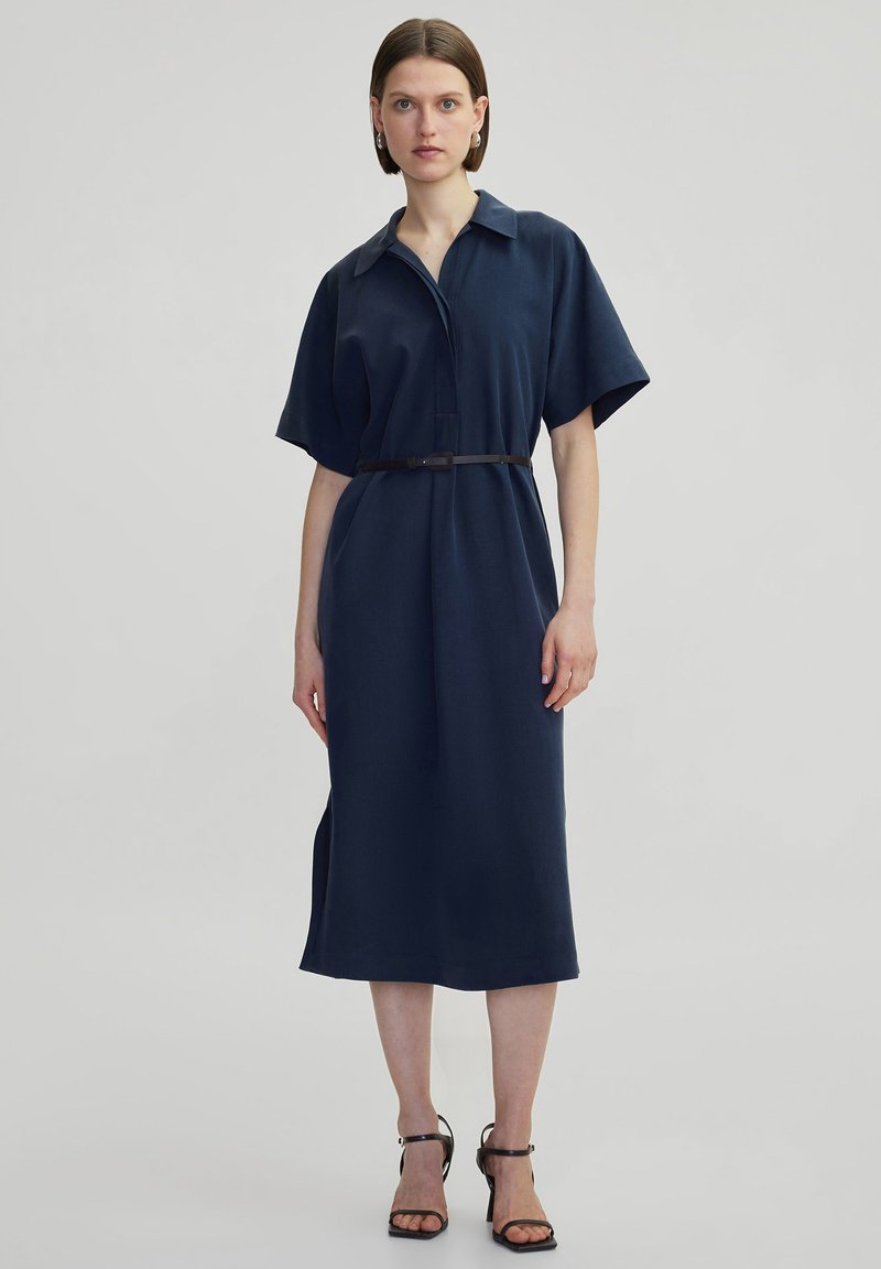 Abito blu navy con cintura, colletto, maniche corte e taglio dritto. Il tessuto appare liscio e presenta delle fessure laterali per facilitare i movimenti.