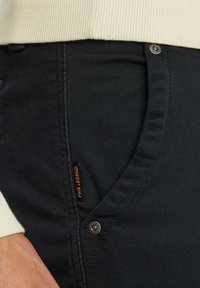Dunkelgraue Hosen mit Seitentaschen, die eine glatte Oberfläche aufweisen und mit einem orangen Etikett versehen sind. Enthält eine metallene Druckknopf-Details.