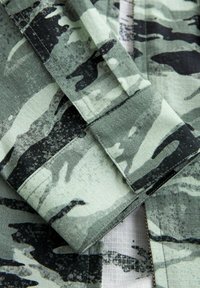 Zeleni i crni kamuflažni materijal s teksturiranom površinom, koji sadrži preklapajuće komade s nepravilan obrascima i detaljima šivanja.