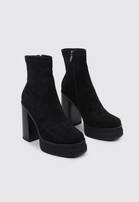 Botas de tobillo de ante negro con un alto tacón de bloque, punta redonda y cremallera lateral. Presentan una textura suave y un diseño minimalista.