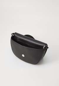 Borsa a tracolla in pelle nera con bordi arrotondati, chiusura a patta con bottone argentato e tracolla regolabile su sfondo bianco.